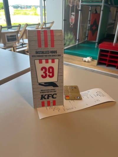 The image shows a table in a KFC restaurant, as indicated by the table sign with the KFC logo and instructions to place the sign visibly for table service. The table number is 39. A receipt and a gold credit card are lying on the table. The receipt shows an order total of €14.65, placed at 7:19 PM on June 13, 2026. The restaurant's phone number is listed as 04 77 31 42 55. The presence of the receipt, credit card, and table sign suggests a customer has recently paid for their meal. Through the window, the location appears to be in Andrézieux-Bouthéon, France. Chairs are seen through the window. Additional tables and booths can be seen in the background of the restaurant, with an object resembling a cat in the background.