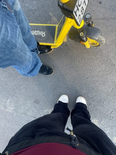 An aerial view captures two individuals standing on a gray asphalt surface, accompanied by a yellow electric scooter. The individual on the left is wearing blue jeans and black shoes, positioned on the scooter. The person on the right is wearing black pants and white shoes. The scooter displays the Russian lettering "Яндекс" and a license plate reading "HC 287."