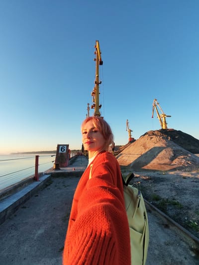 A woman with short, pink hair is taking a selfie on a dock in Bol'shie Mkhi, Russia. She is wearing an orange sweater and a light yellow backpack. In the background, there are several large cranes and piles of gravel. The sun is low in the sky, casting long shadows and creating a warm, golden light. The water of the river is calm and reflects the sky. A red buoy with the number "6" on it is visible on the dock. The scene appears to be a port or industrial area.