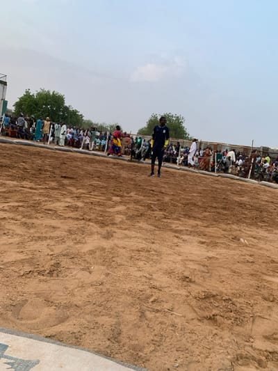 The image shows a sandy outdoor space with a crowd of people gathered along the edge, with one person standing alone in the center of the space. The coordinates (latitude: 13.049, longitude: 5.238) place the location in the city of Sokoto, Nigeria. The person standing alone is wearing a blue shirt with yellow trim and black pants. The crowd of people is comprised of both men and women, mostly wearing traditional clothing. The ground is made of dirt. There is a white plastic barrier in the background and a large gathering of people. There are many trees in the distance behind the people.