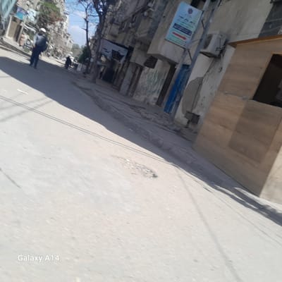 The image shows a street in Nuseirat, Palestinian Territory. A man in dark clothing and a white head covering is walking away from the camera on the left side of the street. In the background, a second person is walking in the same direction. Buildings line the right side of the street. A wooden structure is on the right side. A blue sign with Arabic writing is mounted on a building. The street is paved and appears somewhat dirty. The sky is visible in the distance. The image is slightly blurred, and the camera is tilted.