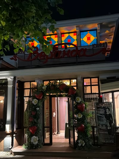 The image shows the entrance to a cafe named "Veranda" in Kislovodsk, Russia. The cafe has a distinctive red sign with white lettering and is decorated with a floral arch featuring large red hearts and white flowers. Above the entrance, there are stained-glass windows with geometric patterns in shades of blue, yellow, and red. To the right of the entrance, a poster displays a woman in black attire and text in Russian, advertising live music, a saxophone, and popular songs. The scene is lit by artificial lights, indicating it is nighttime. The overall impression is of a welcoming establishment, possibly decorated for a festive occasion.