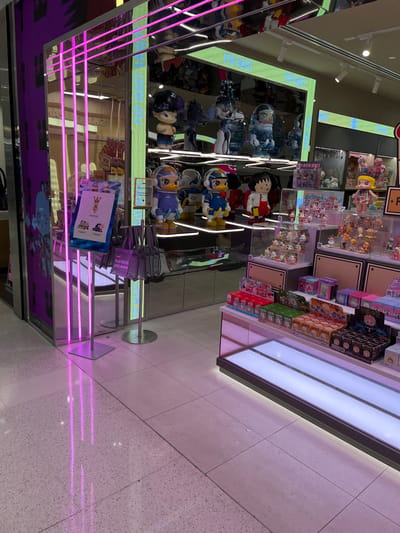This is an interior photograph of a brightly lit retail store, likely specializing in collectible toys or character merchandise. The image is oriented 90 degrees clockwise, showing the floor on the right and ceiling on the left.

The scene features multiple display units filled with various products. On the upper shelves, numerous small figurines are arranged in clear cases and individual boxes, with "POP MART" branding visible on some packaging. Below these, larger, plush-like character figures are displayed on reflective platforms; these include figures in blue and white astronaut-style suits and one in a red and white outfit.

The store utilizes extensive reflective surfaces, possibly mirrored walls or polished metal, which create multiple reflections of the merchandise and lighting. Bright white LED strip lights illuminate the product shelves, while vibrant pink/purple LED lights line a lower display unit, casting colorful reflections onto the light-colored, speckled tiled floor.

A metal queue stanchion with a gray rope is present in the lower center of the corrected image, suggesting a checkout or waiting area. Adjacent to it, a stand holds brochures or flyers, one of which prominently displays "PLAY WITH" and character illustrations. Faint, distorted text like "PLAY NOW" is also visible as a reflection on the mirrored surfaces. A small sign on a display reads "PLEASE KEEP" (likely part of "PLEASE KEEP HANDS OFF").

No people are visible, indicating the shot captures the store's arrangement rather than an active event. The environment is modern and clean, designed to attract customers to the displayed items. The location context provided is Indooroopilly, Australia, though no specific visual cues within the image confirm this.