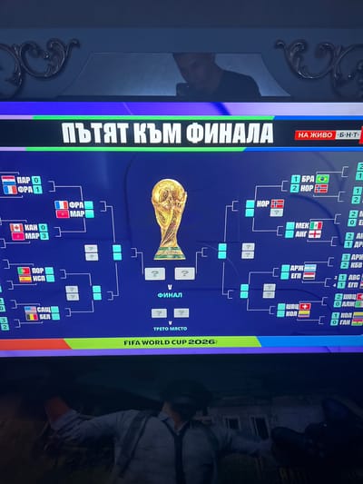 The image shows a television screen displaying a FIFA World Cup tournament bracket. The text on the screen is in Bulgarian and translates to "The Path to the Final". The bracket shows various matches, team names, and scores, with the World Cup trophy at the center. The FIFA World Cup 2026 is also mentioned. The screen is angled and reflects some ambient light, including a portion of a person's face and a decorative wall molding. The bottom of the image is obscured by a dark, blurry area that appears to be the lower part of the television or a surface in front of it, possibly with a person's arm and hand visible. There is no visible indication of Ognianovo, Bulgaria in the image itself.