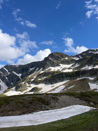 This is a landscape photograph taken in Panichishte, Bulgaria, featuring a dramatic mountain scene. The image is oriented vertically, with the sky occupying the left portion and the mountain range dominating the right.
The sky is a vibrant blue, dotted with fluffy white cumulus clouds. The bright sky suggests it is daytime, likely late morning or afternoon, with clear and fair weather.
The mountain face is rugged and steep, composed of dark, rocky slopes interspersed with patches of snow. The snow is unevenly distributed, clinging to crevices and gullies, indicating a recent thaw or a region with persistent snow cover. Patches of green grass and moss are visible on some of the less steep, exposed areas of the mountain, adding color to the otherwise stark landscape. The texture of the rock is rough and uneven, with visible stratification in places. The snow appears somewhat melted and textured, suggesting it is not fresh snowfall.
The overall scene conveys a sense of raw, natural beauty and the power of the alpine environment. There are no people or man-made objects visible in the frame, emphasizing the wildness of the location. The composition draws the viewer's eye from the expansive sky down to the textured mountain face, highlighting the grandeur of the natural landscape. There is no visible text within the image.