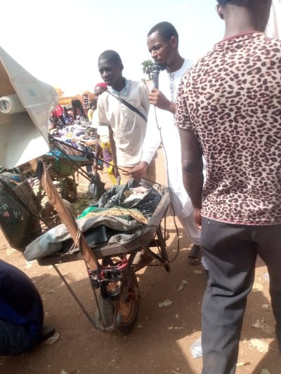 The scene depicts an outdoor gathering in an unpaved area of Kpak, Nigeria, under bright daylight. In the foreground, a dark-colored wheelbarrow, containing various items including patterned textiles, black bags, and electronic components secured with red and black straps, is centrally positioned. A large, light-grey horn speaker is partially visible on the far left, mounted on an unseen structure.
Three male subjects are prominently featured. A young man, wearing a light-colored polo shirt and a dark shoulder strap, stands behind the wheelbarrow, holding a dark cord or wire. To his right, a bearded man, dressed in a white traditional garment, holds a black microphone to his mouth. To the right of the man with the microphone, another male subject stands with his back largely to the camera, wearing a brown and black leopard-print t-shirt and dark trousers. Partially visible in the extreme bottom left foreground is a person's lower leg and foot, clad in dark blue fabric and a dark shoe.
The background reveals an open-air market environment with several indistinct figures, including one wearing a red head covering and yellow clothing. Piles of merchandise, primarily clothing, are visible on the ground or low structures. A yellow sign displaying "POS" is partially legible in the mid-background, amidst the dusty, unpaved terrain.