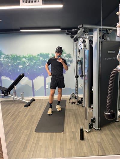 A man in a black shirt, shorts, beanie, and sneakers stands on a black mat in a gym setting. He holds a phone up, seemingly taking a picture in the mirror. Behind him, a mural of palm trees and mountains adorns the wall. A weight machine with cables and a pull-down bar is positioned overhead. An exercise bench sits nearby, along with a water bottle on the floor. The floor is tiled with a light gray wood grain pattern.