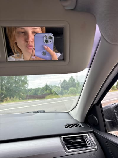 A young woman with blonde hair is reflected in the visor mirror of a car. She is holding a purple smartphone, which is positioned to capture her reflection. She is making a kissing face, with her lips pursed. Her nails are painted a light purple color. The background visible in the mirror reflection shows a road and trees, suggesting the car is in a rural or suburban area. The car's interior is visible in the foreground, including the dashboard and a speaker. The location is identified as Bolshoye Selo, Russia.