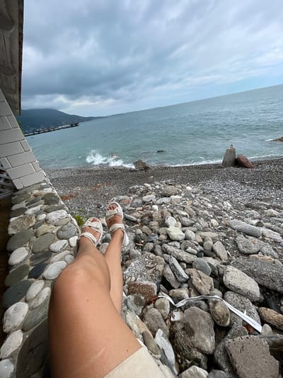 The image shows a person's legs crossed at the knees, wearing white sandals. The person is sitting on a wall made of rounded stones. In the background, there is a pebble beach with a stormy sea and a cloudy sky. A pier with boats can be seen in the distance. The location is the city of Abaata, Abkhazia. The overall scene suggests a seaside relaxation on a gloomy day.