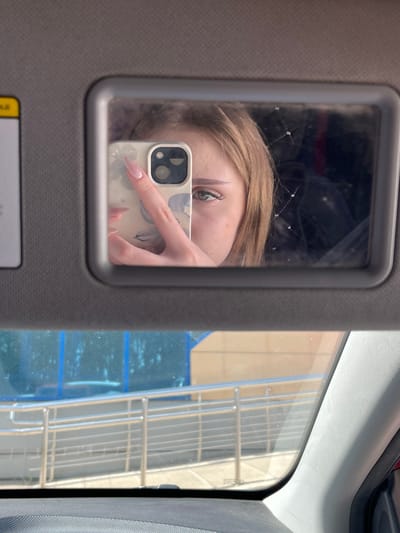 The image is a selfie taken in a car's visor mirror. The reflection shows a young woman holding a smartphone. Her face is partially visible, with one eye and a section of her forehead and cheek in focus. She has blonde hair and is wearing makeup, including false eyelashes. Her right hand is visible, holding the phone, and she has long, light pink fingernails. The background of the mirror reflection is dark and indistinct, with some faint linear patterns suggesting a textured surface or reflections. Below the visor mirror, the car's windshield shows a reflection of the exterior environment, featuring a building with blue accents, metal railings, and a walkway. The car interior is visible at the bottom of the image, including the dashboard and a portion of the A-pillar. The image appears to be taken during the daytime, with sunlight casting shadows and highlighting textures.