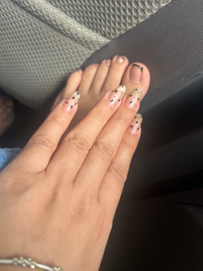 The image displays a close-up view of a person's hands and feet, focusing on their manicured nails. The nails on the hands are long and oval-shaped, adorned with a translucent pink polish and black polka dots. The toenails also feature a similar design, with some having a French tip with black accents, while others have the black polka dot pattern. The feet and hands appear to be resting on a textured, gray surface, possibly upholstery or a car seat, with a dark, out-of-focus background. There are no discernible elements within the image that indicate the specific location in Conuco Viejo, Venezuela, or any urban environmental features. The scene is devoid of any people other than the implied presence of the hand and foot owner, and there are no active interactions or events occurring.