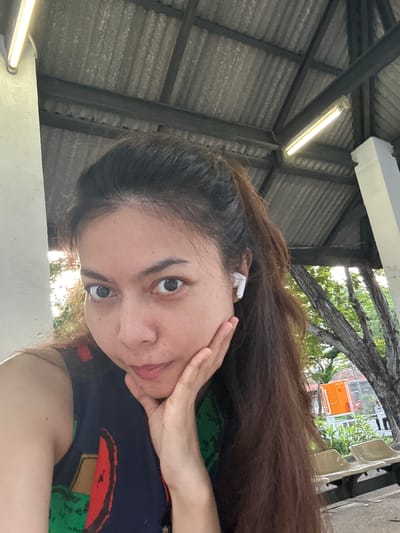 A woman with long, dark brown hair pulled into a ponytail, wearing a dark blue sleeveless top with a colorful pattern, is taking a selfie. She is resting her right hand on her cheek. She is wearing a white wireless earbud in her right ear. The background appears to be an outdoor covered area with a corrugated metal roof supported by beams and pillars. Fluorescent lights are visible on the ceiling. Outside the covered area, trees and some outdoor seating are visible. The scene suggests a public area like a bus stop or waiting area, possibly in Tha Khlong Town Municipality, Thailand, given the vegetation and architecture typical of the region.