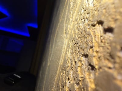 The scene is an interior environment located in Bukovlak, Bulgaria. The foreground is dominated by a highly textured, granular surface, appearing predominantly gold or bronze in color, extending from the bottom right to the top right of the frame. This surface exhibits irregular relief and reflective qualities, suggesting a metallic or heavily painted finish. Towards the center-left, a smooth, light-colored vertical element, possibly a wall or partition, separates the textured foreground from a darker background. The background is dimly lit, featuring distinct angular patterns of bright blue light, indicative of an artificial illumination source such as LED strips. In the lower-left section of the background, a dark surface, possibly a piece of furniture, is visible, upon which rests a small, black electronic object, consistent with a remote control, showing a white accent. No individuals are present in the image.