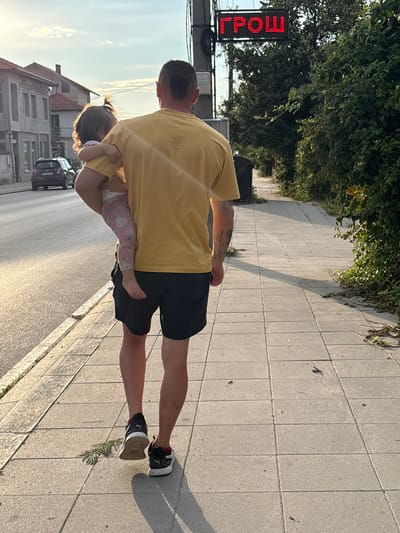 A man wearing a yellow t-shirt and black shorts carries a young child in his left arm. The man is walking on a sidewalk next to a street. The child is facing away from the camera and is wearing a pink outfit. In the background, there are buildings, trees, and a digital sign that displays red letters. The image appears to be taken during the daytime, possibly in the late afternoon, given the lighting. The sign in the background displays the word "ГРОШ" in Cyrillic, which translates to "GROSH" in English. This is likely a street name or a business name in Oreshak, Bulgaria. The sidewalk is made of concrete tiles, and there is some debris on the ground. A dark-colored car is visible on the street in the distance.