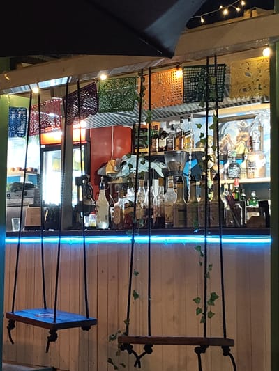 The image depicts a bar with two swings in the foreground. Above the bar, colorful, patterned paper decorations are strung up. Various bottles and objects line the bar counter. In the background, a large painting is visible on the wall. A blue light illuminates the edge of the bar. The bar is located in Rotes, Spain. No people are visible in the image.