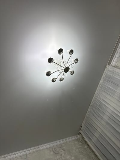 A low-angle shot captures a chandelier with eight arms extending from a central base, each arm ending in a decorative light fixture. The chandelier is positioned against a pale, possibly stretch, ceiling, with a bright light source emanating from above and behind it, creating a halo effect. On the right side of the image, a white, sheer curtain is visible, hanging from a curtain rod, partially obscuring a patterned wallpaper. The ceiling features decorative molding along the top edge of the wallpaper. The image appears to be taken indoors, likely in a residential setting, in the city of Zhanaozen, Kazakhstan. There are no people or animals visible in the frame, and the scene is static.