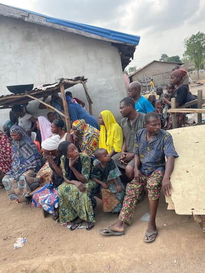 The image depicts a daytime outdoor gathering of approximately 20-25 individuals, including adult men, women, and children, in a residential area of Osara Gada, Nigeria.
The foreground and mid-ground show a crowd predominantly seated on low wooden benches or directly on the dry, uneven dirt ground. Most individuals are oriented towards the left side of the frame, suggesting they are observing an event or person outside the immediate view. Attire consists primarily of traditional patterned fabrics; many women wear head coverings in various colors and patterns (e.g., yellow, blue, multi-colored, white). A man in the front right wears a blue patterned shirt, pink and green patterned trousers, and grey sandals. A woman in the mid-ground, left of center, wears a white head covering and a dark top, with her chin resting on her hand. Adjacent to her is a young boy in green patterned clothing. A woman further to the right holds a small child.
In the background, a single-story building with a light grey, rough-plastered exterior wall and a partially damaged blue corrugated metal roof serves as a backdrop. Faint white writing is visible on the wall. A makeshift lean-to shelter, constructed from wooden poles and corrugated metal sheets, extends from this building, with a dark, empty basin resting on its roof. Further in the distance, other simple buildings with corrugated metal roofs are visible. The ground is dry, dusty earth, with a piece of discarded paper or cardboard visible in the immediate foreground. The sky is overcast.