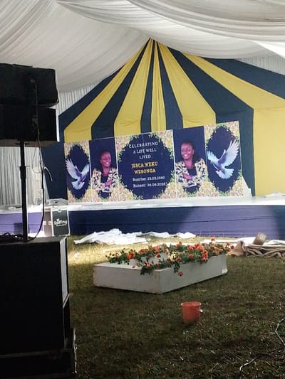 An outdoor event tent, located in Marenyo-Shianda ward, Kenya, is depicted. The tent features a white draped ceiling and a blue and yellow striped fabric backdrop at the rear. A dark blue stage is positioned in front of this backdrop. Three large memorial banners are displayed on the stage. The central banner reads "CELEBRATING A LIFE WELL LIVED JESCA WEKU WEBONGA Sunrise: 23.03.1980 Sunset: 26.08.2023," featuring a smiling woman's portrait framed by floral designs. Two identical side banners display the same woman's portrait, floral designs, and a white dove in flight.
In the mid-ground, on grassy turf, a white rectangular planter filled with numerous small orange-pink flowers is present, positioned horizontally. An orange plastic bucket rests on the grass behind this planter. To the left foreground, two large black professional audio speakers are stacked on a stand. White tarpaulins or fabric pieces are loosely scattered on the grass in front of the stage. Brown, coarse fabric is visible on the far right of the grassy area. No individuals are present in the scene.
