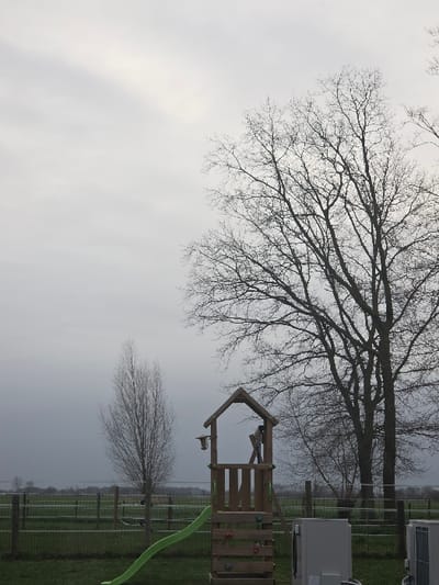 The image displays an outdoor scene in Eeklo, Belgium, under an overcast, light grey sky. In the immediate foreground, green grass covers the ground. Centrally positioned is a wooden children's play structure, featuring a peaked roof, a green plastic slide extending downwards to the left, and multi-colored climbing holds attached to its side panel. A dark, tubular object, possibly a periscope or telescope, is mounted on the play tower's platform, and a small, light-colored object hangs from the left side of its roof. To the far right, two light-colored, rectangular outdoor units, consistent with air conditioning condensers or heat pumps, are situated on the grass. A large, bare deciduous tree with extensive branching dominates the right side of the frame. A smaller, slender bare deciduous tree stands towards the mid-left. A multi-strand wire fence with wooden posts runs horizontally across the mid-ground, demarcating the foreground from a flat, green field or pasture. Distant landscape features and trees are visible on the horizon beneath the grey sky. No people, animals, or active subjects are present. The lack of foliage on the trees indicates a dormant season.