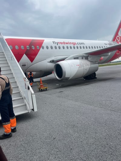 An airplane with the livery "flyredwings.com" is parked on the tarmac under a cloudy sky. A set of mobile stairs is positioned at the side of the plane. Two individuals are visible near the stairs: one is standing on the ground wearing dark overalls and bright orange leg coverings, and another person in similar attire is partially visible on the stairs. Another worker in an orange vest and dark pants is near the plane's engine. A yellow cart with an orange traffic cone is positioned on the tarmac. The aircraft's registration number, RA-89157, is visible on the fuselage. The environment appears to be an airport runway or apron.