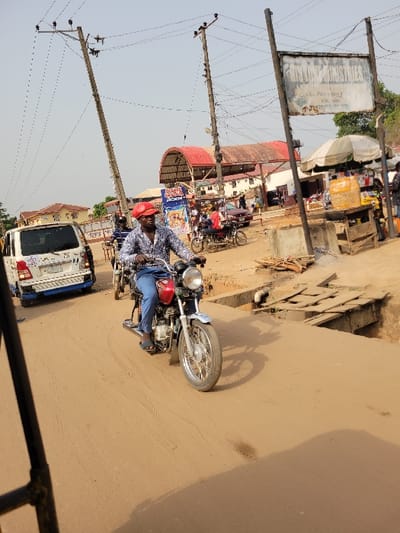 The image depicts a street scene in the city of Puerto López, Colombia, under a hazy sky.
In the foreground, a dark-skinned adult male is operating a red and silver motorcycle, traveling from left to right. He is wearing a red cap, a patterned long-sleeved shirt (blue, white, black), blue trousers, and dark sandals. The motorcycle features a red fuel tank, silver fenders, a chrome headlight, and spoked wheels. Shadows from the motorcycle and an unseen object (likely the photographer's vehicle) are cast upon the unpaved, light brown road surface, which shows signs of dust and unevenness.
To the immediate right of the motorcycle, a raised concrete structure with wooden planks spans a ditch, serving as a culvert or small bridge. Further right, a market stall with a light-colored umbrella displays various packaged goods and bottles. A large, weathered rectangular sign, mounted on two poles, is partially visible above the market stall, bearing the faded text "ANYAM INDUSTRIES."
On the left side of the road, a white van with a blue stripe on its lower body is parked, facing away from the camera. Utility infrastructure is prominent, with two leaning wooden poles supporting multiple overhead power lines stretching across the street.
In the mid-ground, other individuals and vehicles are visible. Behind the primary motorcyclist, additional motorcycles are present. Further back, a building with a distinctive red, arched roof is visible, along with other low-rise structures. Figures can be seen near the market stall and in the vicinity of the red-roofed building, including one person on a motorcycle wearing a red shirt. The overall environment suggests a bustling, commercial street with a mix of permanent and temporary structures and an unpaved road.