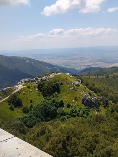 This is a wide, high-angle shot of a mountainous landscape, likely taken from a viewpoint or observation deck. In the foreground, a concrete platform with some scattered coins occupies the lower left corner of the frame. The middle ground features a rocky, grassy ridge with scattered green trees and shrubs. A dirt path winds along the ridge, and several small figures, likely hikers, are visible on or near the path. One figure stands near a cross at the highest point of the ridge.
The background reveals a vast valley with rolling hills covered in dense green forests, receding into the distance. The far distance is hazy with a flat plain dotted with agricultural fields and a small town. The sky above is a clear blue, with scattered fluffy white clouds. The scene depicts a sunny day in a natural environment, with a clear view of the surrounding terrain.
This location is very likely the Buzludzha monument, which is located near the city of Shipka, Bulgaria. The monument is an abandoned, Brutalist structure that was once a meeting place for the Bulgarian Communist Party. It is situated on a mountain peak, offering panoramic views of the surrounding countryside, consistent with the landscape depicted.