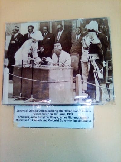 The image displays a physical photograph of a historical black and white scene, mounted on a light-colored wall, with a descriptive label beneath it. This displayed image is located in South Sakwa ward, Kenya.
The historical photograph depicts eight men participating in a formal signing ceremony. In the foreground, a dark-skinned man identified as Jaramogi Oginga Odinga, wearing a light-colored suit, tie, and a distinct patterned cap, is seated at a rectangular wooden desk. He is actively signing a document with a pen in his right hand. To his immediate left (viewer's right), a light-skinned man in a white pith helmet and a light-colored suit leans forward over the desk, observing the signing. To Odinga's right (viewer's left of the central group), a man identified as Colonial Governor Ian McDonald stands stiffly, dressed in a dark ceremonial uniform with a white plumed hat, epaulets, a prominent sash, and a ceremonial sword or scabbard at his hip. Behind these central figures, five additional dark-skinned men in dark suits and ties are standing and observing the proceedings. These individuals are identified as Jomo Kenyatta, Mboya, James Gichuru, Joseph Murumbi, and J.D. Ouende.
On the wooden desk, documents and at least two microphone stands are visible. In the immediate foreground, a rope barrier supported by dark metal poles delineates the ceremonial area. The background of the scene appears to be an interior or covered outdoor space, possibly a wall or window structure.
Beneath the photograph, a white rectangular label provides textual information: "Jaramogi Oginga Odinga signing after being sworn in as a new minister on 13th June, 1963. From left, Jomo Kenyatta, Mboya, James Gichuru, Joseph Murumbi, J.D. Ouende and Colonial Governor Ian McDonald."