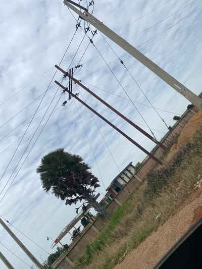 The image depicts a roadside view in Kuru, Nigeria, characterized by a cloudy sky and a tilted perspective. Prominent in the foreground are multiple utility poles with overhead electrical wires, some made of concrete and others of weathered metal. A large palm tree with dense, fan-shaped fronds stands to the left, its trunk partially obscured. Behind the tree, a low-rise building with a light-colored facade and darker window frames is visible, situated behind a beige-colored wall. Adjacent to the building, there is an area with dry, tall grass and scattered debris. To the right of the scene, a pile of light brown material, possibly soil or sand, is stacked. A portion of a gas station canopy, identified by red and white signage including the text "OIL & GAS LTD", is visible beneath the palm tree. The ground appears to be a mix of dirt, gravel, and sparse vegetation. The overall impression is that of a semi-urban or rural roadside environment under an overcast sky.