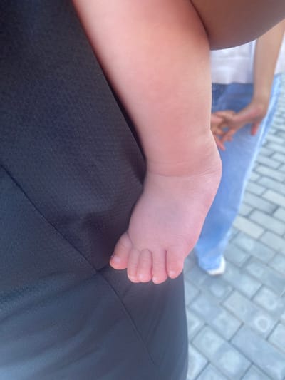 A close-up, waist-level shot shows the bare foot of a baby resting against the dark fabric of an adult's clothing. The baby's skin is pink and soft, with clearly defined toes and fingernails. The adult's clothing appears to be made of a textured material, possibly athletic wear. In the background, out of focus, another person is visible wearing a white top and blue jeans, standing on a paved surface. The paving stones suggest an outdoor urban setting, potentially in Manas, Kyrgyzstan, though this location is not explicitly confirmed by visual cues within the image.