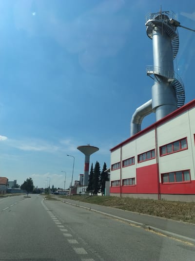 The image depicts a view of an industrial area in Horažďovice, Czechia, under a clear blue sky with scattered clouds. In the foreground, a paved road curves to the left, with dashed white lines marking the lanes. To the right, a modern industrial building with white and red panels and red-framed windows is visible. Adjacent to this building, a tall, cylindrical metal structure with a spiral staircase ascending its exterior stands prominently against the sky. Further in the background, a water tower with a red and white cylindrical base and a wider, bowl-shaped top is observable, along with several tall evergreen trees and other smaller buildings. The overall scene suggests an industrial or commercial zone within the city.
