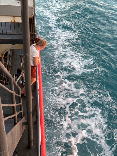 The image shows a woman standing on the deck of a ferry in Nathon, Thailand, looking at the ocean wake. She is wearing a white t-shirt and red shorts. A man in a yellow vest is visible on the lower deck behind her. The ferry is moving, creating a foamy wake in the turquoise water. The sun is setting, casting a warm light on the scene.