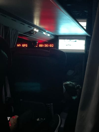 Inside a dark vehicle, a woman with a bun hairstyle sits facing forward. A "NO GPS" alert and a digital timer display "00:36:02" are visible on a console. A window or opening emits bright light. A portion of the vehicle's interior appears on the left side, illuminated by red light. The overall scene is dimly lit, obscuring many details.
