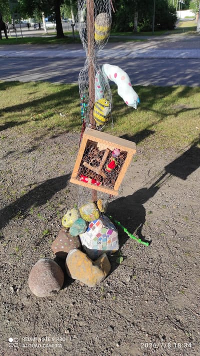 The image shows a decorative structure made of natural and craft materials, likely an art installation, situated outdoors in a public area with a paved road and grassy verge visible in the background. The structure features a wooden box filled with pinecones, possibly a shelter or feeder for small creatures. Surrounding the base of the structure are several decorated rocks, one of which is covered in colorful mosaic tiles. Suspended above the box by wire mesh are several fabric-like objects, including a yellow and black striped one resembling a bee, and a white object with pink polka dots. The scene is illuminated by sunlight, casting shadows on the ground. The timestamp in the lower right corner indicates the image was taken on July 8, 2026, at 18:34. The presence of streetlights, trees, and a road suggests an urban or suburban environment. Based on the style and context, this installation may be part of a public art initiative or a local community project in Iisalmi, Finland, potentially located in a park, along a walking path, or near a community center.