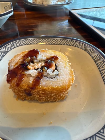The scene depicts a single, deep-fried sushi roll served on a light-colored ceramic plate in an indoor dining establishment in Altenheim, Germany. The sushi roll, positioned centrally, is approximately cylindrical, with a golden-brown, crispy breaded exterior. Its cross-section reveals layers of white rice, dark nori, and a filling that includes light pink/orange material, consistent with salmon or similar seafood. A thick, dark brown, glossy sauce is generously drizzled over the top of the roll. The plate features a wide rim decorated with a repeating blue and white geometric chevron pattern. The surface beneath the plate is a dark brown wooden table with visible grain. In the blurred background, to the left, a portion of a small, white ceramic bowl with a dark patterned rim is visible. To the right, a dark-cased electronic device, possibly a smartphone or tablet, rests on the table, displaying a reflection. Another indistinct plate with light-colored food is partially visible further in the background. No individuals are visible, indicating the food is prepared for consumption.