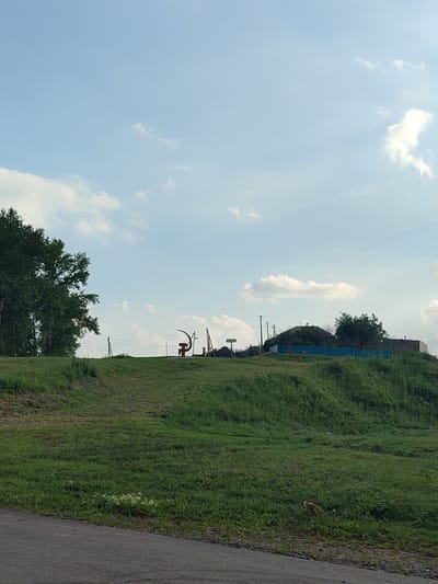 An outdoor, daytime shot captures a grassy hill under a partly cloudy sky. On the crest of the hill, a stylized sculpture resembling a sickle and hammer stands as a prominent landmark. In the distance, a few houses and utility poles are visible. A large green tree is situated on the left side of the frame. The foreground features a dark asphalt road. The scene is likely located in the village of Karabashyevo, Russia, given the presence of a sickle and hammer sculpture, which is a common symbol in rural Russia.