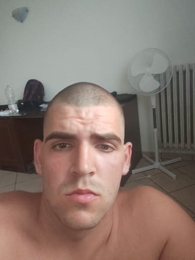 This media file is a close-up selfie of a man with a shaved head. He is facing the camera, and his expression is neutral. The background appears to be an indoor setting, possibly a bedroom, with a desk, a fan, and a radiator visible.
The man's face is the primary focus. He has light brown eyes, dark eyebrows, and a slight stubble on his chin. He is shirtless, and his bare shoulders are visible.
In the background, to the left of the man, a desk with some items on it is partially visible. These items include a clear plastic water bottle and what appears to be a backpack or bag with a patterned design.
To the right of the man, a white pedestal fan stands next to a white radiator. The fan has three blades and a protective grill. The radiator is made of vertical metal bars.
A light fixture is visible on the wall in the upper left corner of the image.
The overall lighting suggests it is daytime, but there are no clear indicators of the time of day or weather conditions outside. The setting appears to be a residential interior.
There is no visible text in the image. The dominant emotion conveyed by the subject's expression is neutrality. The location context provided is Guarene, Italy.