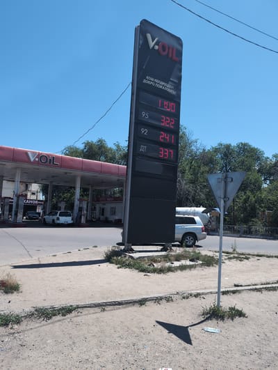 A digital price board displays fuel prices at a V-OIL gas station. The sign shows prices for different octane fuels: 95 at 322, 92 at 241, and "ДТ" (diesel fuel) at 337. Above these, a price of 1100 is displayed without an octane number. Text in a foreign language reads "ҚОШ КЕЛДІҢІЗДЕР! ДОБРО ПОЖАЛОВАТЬ!" which translates to "Welcome!". The gas station has a red roof supported by white columns. Several cars are visible, including a white SUV at a pump and a dark sedan. In the background, trees and a light-colored building are present. A grey triangular sign on a pole stands in the foreground to the right. The ground is composed of dirt and gravel. This scene is located at the V-OIL gas station in Otegen Batyr, Kazakhstan.