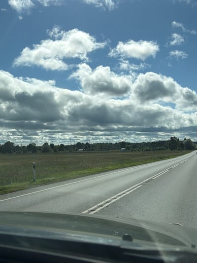 This media file is a photograph taken from inside a vehicle, showing a road and the surrounding landscape. The road, paved with asphalt, stretches into the distance and is marked with white lines. To the left of the road is a grassy verge, and beyond that, a line of trees and vegetation. In the background, several wind turbines are visible. Above the landscape, the sky is filled with a dramatic display of clouds against a blue sky. Some clouds are large and cumulus, while others are thinner and more layered. The lighting suggests it is daytime, likely late morning or afternoon, with bright sunlight illuminating the scene. The overall mood is one of travel and open space, with the weather appearing partly cloudy but pleasant. No people are visible in the image. The scene is likely in a rural or semi-rural area. There is no visible text in the image.