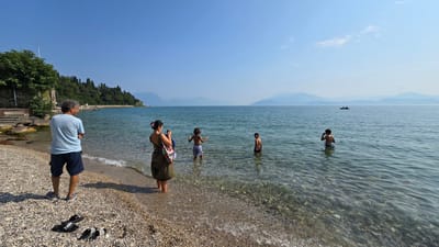 In Sirmione, Italy, on a sunny day, a man with grey curly hair, wearing a light blue t-shirt and dark shorts, stands on a pebble beach facing away from the camera towards a clear blue lake. To his right, a woman with dark hair pulled up, wearing a long olive-green dress and carrying a patterned bag, stands in the shallow water, holding a phone as if to take a picture. Three young boys are in the lake, two further out and one closer to the woman. One boy wears blue and white striped shorts, another wears dark shorts, and a third, further out, is only visible from the shoulders up. In the background, a distant shoreline is lined with trees and hills under a clear sky. Further out on the water, a small boat with two people is visible. On the beach in the foreground, black flip-flops are laid out on the pebbles.