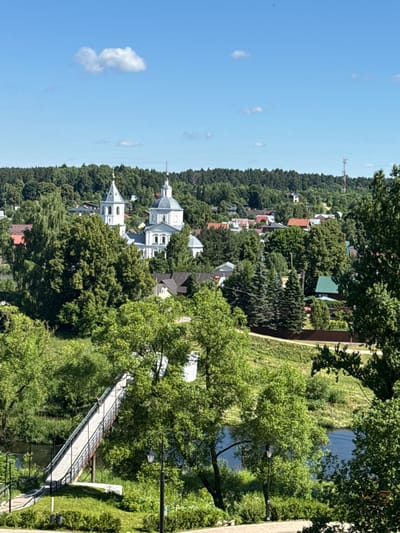 The image presents a serene, elevated view of the city of Vereya, Russia. In the foreground, a pedestrian bridge spans a calm body of water, with lush green trees and manicured bushes lining its approach and banks. A solitary lamppost stands near the bridge's entrance. Beyond the bridge, the landscape transitions into a dense forest that covers rolling hills. Nestled within this verdant expanse are several buildings, most notably a prominent white church with a distinctive bell tower and a large dome, indicative of traditional Russian architecture. The church is surrounded by a cluster of houses with red-tiled roofs, suggesting a residential area. The sky above is a clear, bright blue, punctuated by a few fluffy white clouds. In the far distance, a tall telecommunication tower is visible, hinting at the city's infrastructure. The overall scene conveys a sense of tranquility and offers a picturesque glimpse of Vereya's natural beauty and architectural heritage.