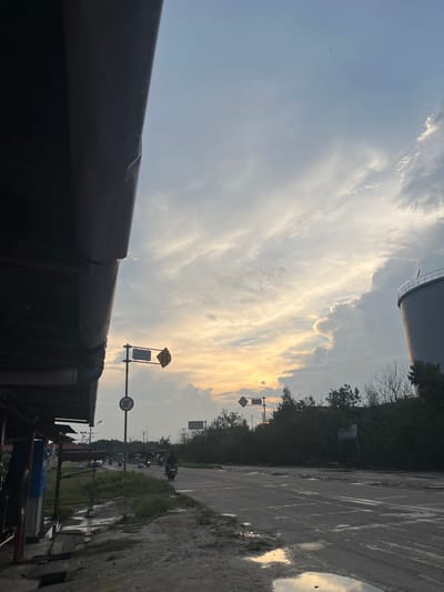 The image depicts a street scene in Dumai City, Indonesia, during what appears to be sunset. The foreground is dominated by the dark, overhanging roof of a structure on the left, possibly a roadside stall or shop. A wet, muddy road stretches into the distance, reflecting the sky. Several motorcycles are visible on the road, with riders appearing to be locals. Traffic signals and road signs, including a speed limit sign indicating 20 km/h, are present. In the background, lush green foliage and trees line the roadside, partially obscuring a large, grey cylindrical industrial tank on the right. The sky is filled with a mix of clouds, with the warm hues of sunset breaking through, creating a dramatic contrast with the overcast conditions. The overall impression is that of a functional, everyday street in a tropical urban environment, perhaps after a recent rain.