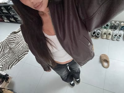 An individual stands indoors on a light grey tiled floor in Tielrode, Belgium, captured from an overhead perspective. The person wears a dark brown zippered jacket, partially unzipped over a white ribbed tank top that exposes a segment of the midriff, dark-wash jeans, and black sneakers with white laces. Long dark hair is visible, and the lower portion of the face, including lips with nude-toned lipstick, is present. A thin silver-colored necklace is also visible. To the individual's right, a single beige furry slipper rests on the floor, while approximately 10-12 pairs of various athletic shoes are arranged in a line against a wall further in the background. To the left, a brown and white zebra-print rug is partially visible on the floor. In the upper left background, a dark multi-compartment shoe rack containing shoes is partially observed, and in the lower left, a wooden crate holds dark fabric items.