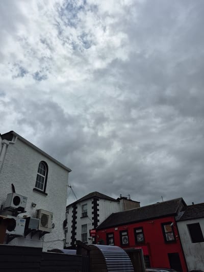 The photograph, taken at an upward angle in Carrick-On-Shannon, Ireland, captures a cluster of buildings under a cloudy sky. On the left, a white building with a textured facade features an arched window and multiple outdoor air conditioning units mounted on its side. To its right, a building with a white facade and black trim around its windows and corners is visible. Further to the right, a vibrant red building with several windows displays signs, one of which reads "APACHE" with "OPEN" underneath. The rooftops of the buildings are tiled. In the foreground, a dark wooden fence runs horizontally across the bottom of the frame, with sections of corrugated metal roofing visible behind it. The sky is filled with a dense, grey cloud cover, suggesting overcast weather. No people are discernible in the image.