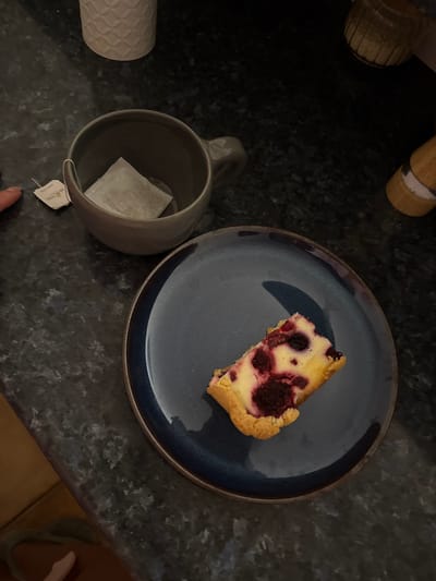 The image shows a top-down view of a dark countertop with several objects. A slice of raspberry cheesecake sits on a blue plate. A gray mug containing a tea bag with the tag visible is to the left of the plate. Further to the left, there is a white patterned container and a wooden object. The countertop surface is dark and speckled. The scene appears to be set indoors. There is no evidence to determine this is the city of Zankovići, Montenegro.