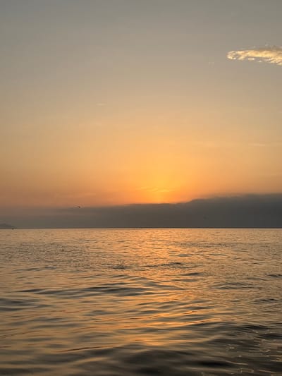 The image depicts a sunset over the ocean, likely taken in or near Ancón, Peru, given the prompt's context. The sky transitions from a light, almost white-gray near the top left to a gradient of orange and yellow toward the horizon, where the sun is setting. The ocean occupies the right half of the image, reflecting the golden sunlight. The water surface is rippled, creating streaks and highlights. A faint silhouette of land is visible on the horizon. A single bird is seen in flight near the horizon line.