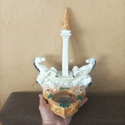 A human hand is holding a decorative souvenir item against a plain, light brown wall in Pasir Muncang, Indonesia. The item, a miniature replica of the Monumen Nasional (Monas), features a tall white column topped with a golden, flame-like finial. The base of the replica is ornate, rendered in gold and light brown, and includes two rearing white horse figures with black accents positioned on either side of the central column. The word "Monas" is inscribed in green script on the front face of the base. A circular, recessed depression, possibly an ashtray, with gold and green decorative elements, is integrated into the lower front section of the base and contains a dark residue. A dark, undefined surface is visible below the hand and object.