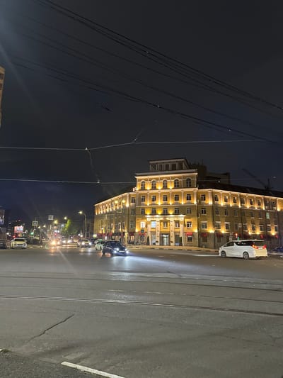 The image shows a street in Komgaron, Russia at night. A large, light-colored building with many windows dominates the right side of the frame. Cars are parked along the street, and traffic is visible. Utility lines crisscross the dark sky on the left side. The street appears wet and reflects the city lights. Tram tracks run along the road. Street lights and storefront signs illuminate the scene.