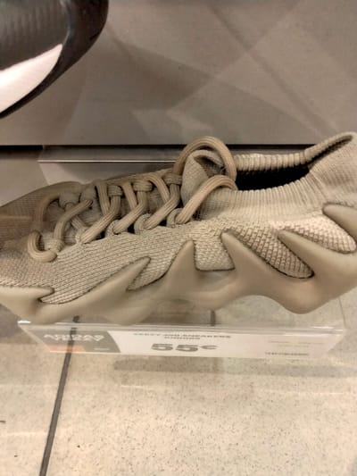A single Yeezy 450 sneaker in a muted earth tone sits on a clear plastic shelf. The sneaker features a knit upper with unconventional lacing and a unique, sculptural midsole. A partially visible tag underneath the shoe advertises "Yeezy 450 Sneakers Juniors" for 55€. A second shoe of different style is partially visible in the upper left corner of the shot. The background and foreground are tiled, adding a sense of a retail environment.
