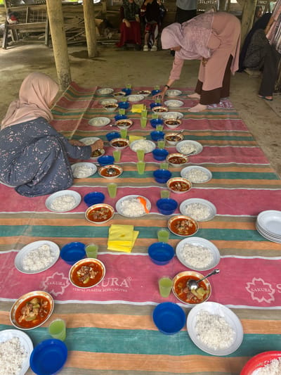 In Pulo, Indonesia, a group of individuals are gathered for a meal on a striped mat spread on a dirt floor, likely outdoors or in an open-air structure. The mat is set with multiple servings of rice in white oval plates, accompanied by bowls of meat stew and glasses of green beverage. Two women, one in a pink hijab and the other in a blue and grey patterned hijab and tunic, are visible near the food, with the woman in pink appearing to serve or adjust the meal. Other people, mostly obscured, are seated in the background. The environment suggests a rustic, communal setting.