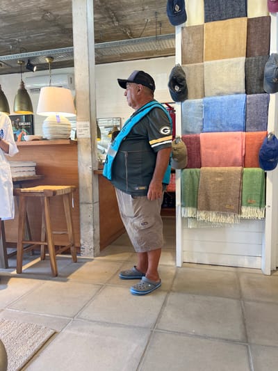 A man in a green and yellow Packers shirt, khaki shorts, and grey Crocs stands in a store. A blue lanyard hangs around his neck. Behind him is a shelf with folded blankets and baseball caps. To the left is a wooden display case with lamps, books, and other items. A wooden chair sits nearby. The floor is tiled.